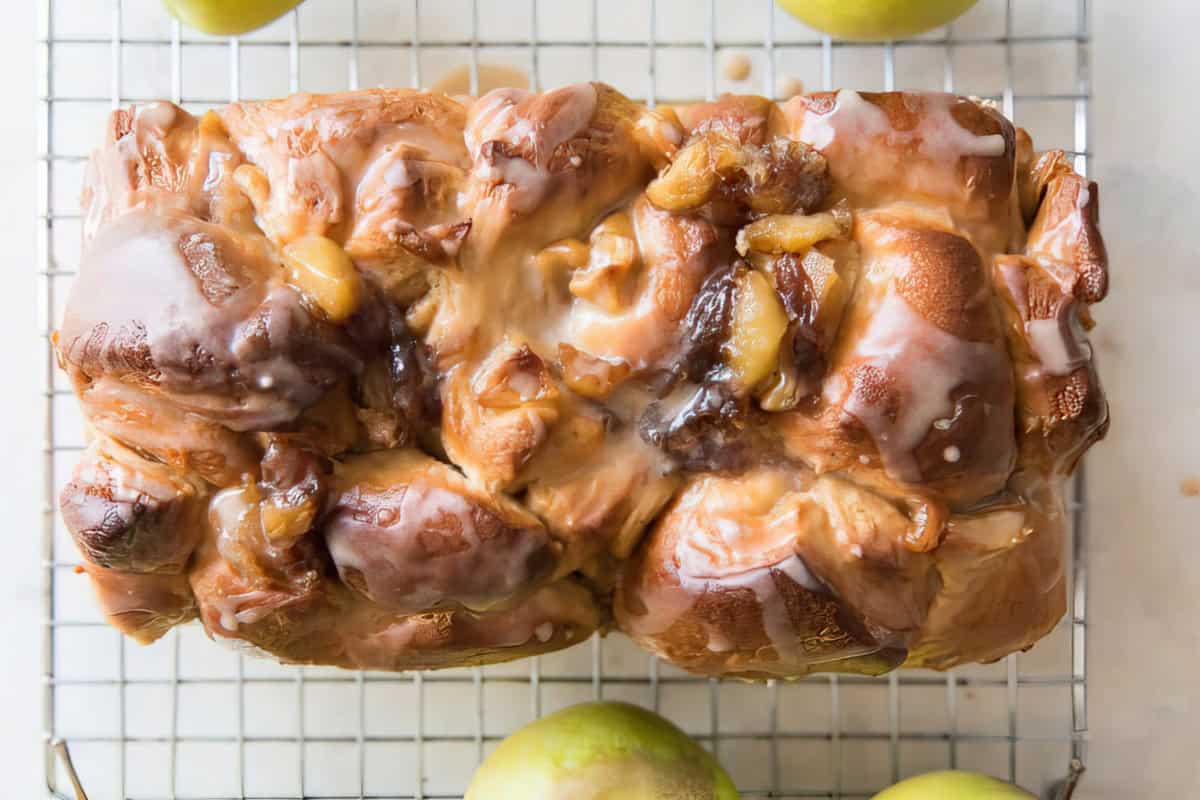 A close-up view of a freshly baked pull-apart apple cinnamon bread with glaze drizzled on top. The loaf is cooling on a wire rack, and a few green apples are partially visible around it. The bread has a golden-brown crust with appealing swirls of cinnamon and apple filling.