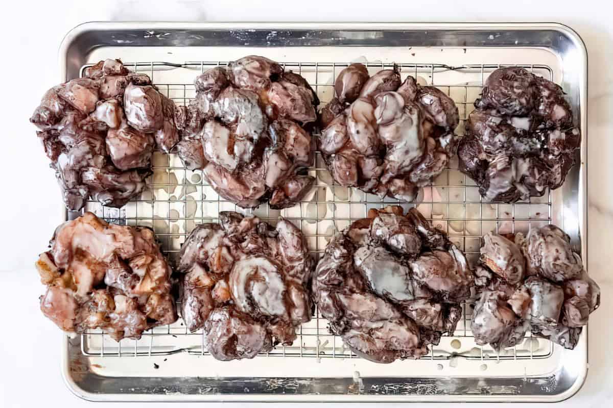 A metal baking tray holds seven chocolate-glazed apple fritters arranged on a wire rack. Each fritter is irregularly shaped, covered in a glossy glaze, and filled with chunks of apples, creating a rustic and delicious appearance.