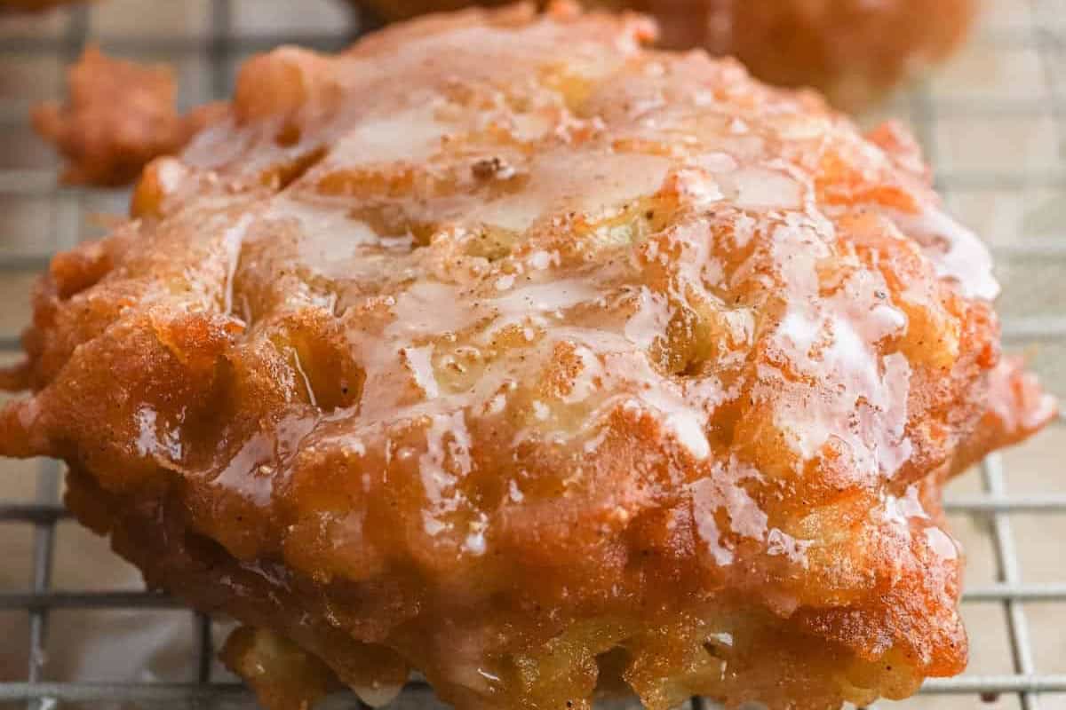 A close-up photo of a glazed apple fritter on a cooling rack. The fritter has a golden brown, crispy exterior with visible chunks of apple, and is drizzled with a shiny glaze.