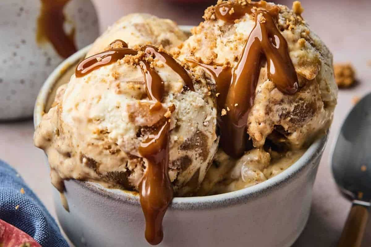 A close-up view of a bowl filled with scoops of caramel swirl ice cream. The ice cream has a rich, creamy texture with visible ribbons of caramel drizzled on top, adding a glossy finish. The bowl is placed on a smooth surface near a spoon and a blue fabric.