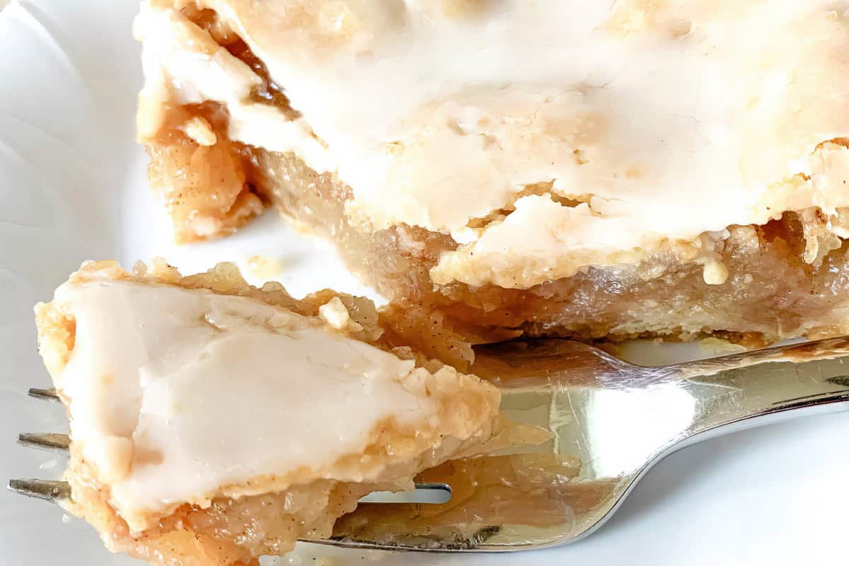 A close-up of a slice of apple pie topped with a thick white glaze. A fork holds a piece of the pie, showing the flaky crust and gooey apple filling underneath. The pie sits on a white plate.