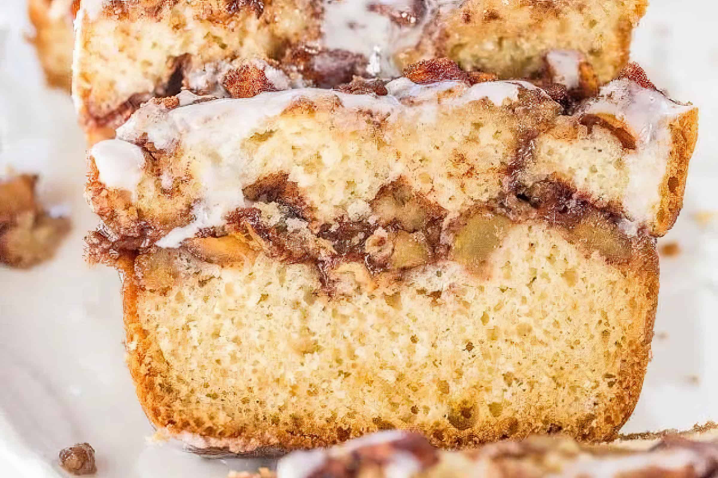 Close-up of a slice of cinnamon swirl quick bread topped with a light drizzle of icing. The bread features distinctive layers of cinnamon filling and nuts, creating a marbled effect throughout the moist, light-colored crumb.
