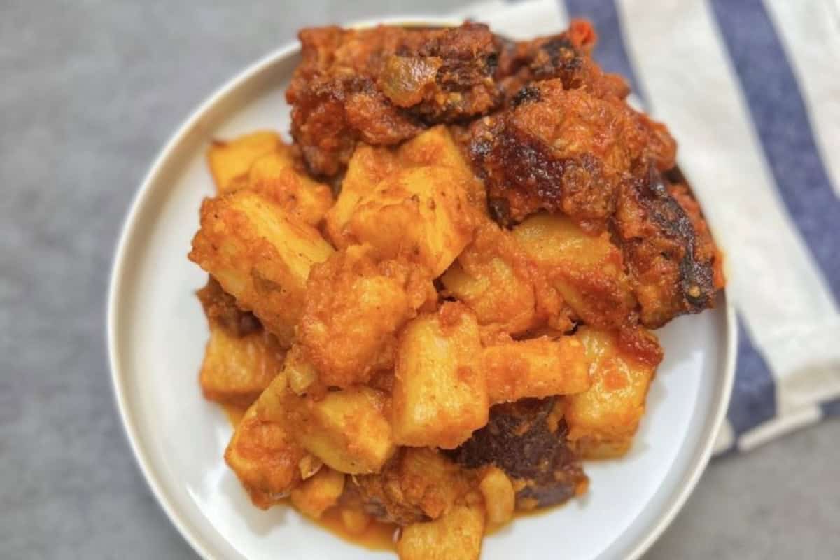A plate of yam porridge topped with rich, seasoned fish pieces. The dish has a reddish-orange hue, indicating a tomato-based sauce. The background includes a folded striped towel on a gray surface.