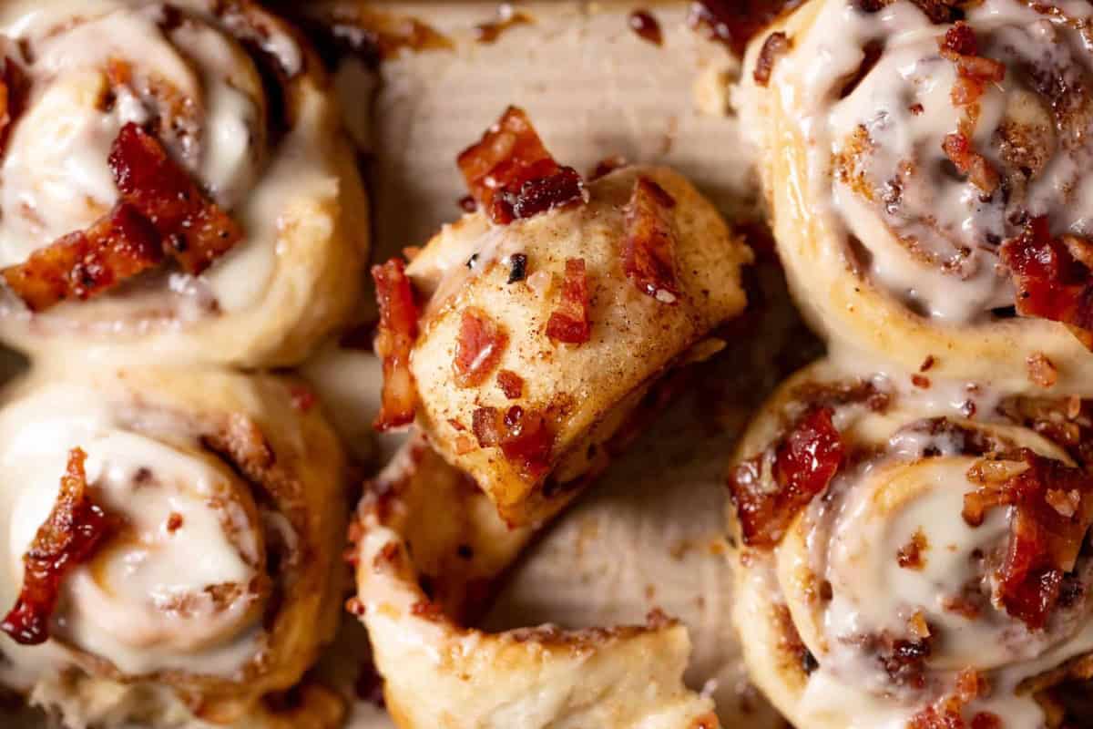 A tray of cinnamon rolls with icing, topped with pieces of bacon.