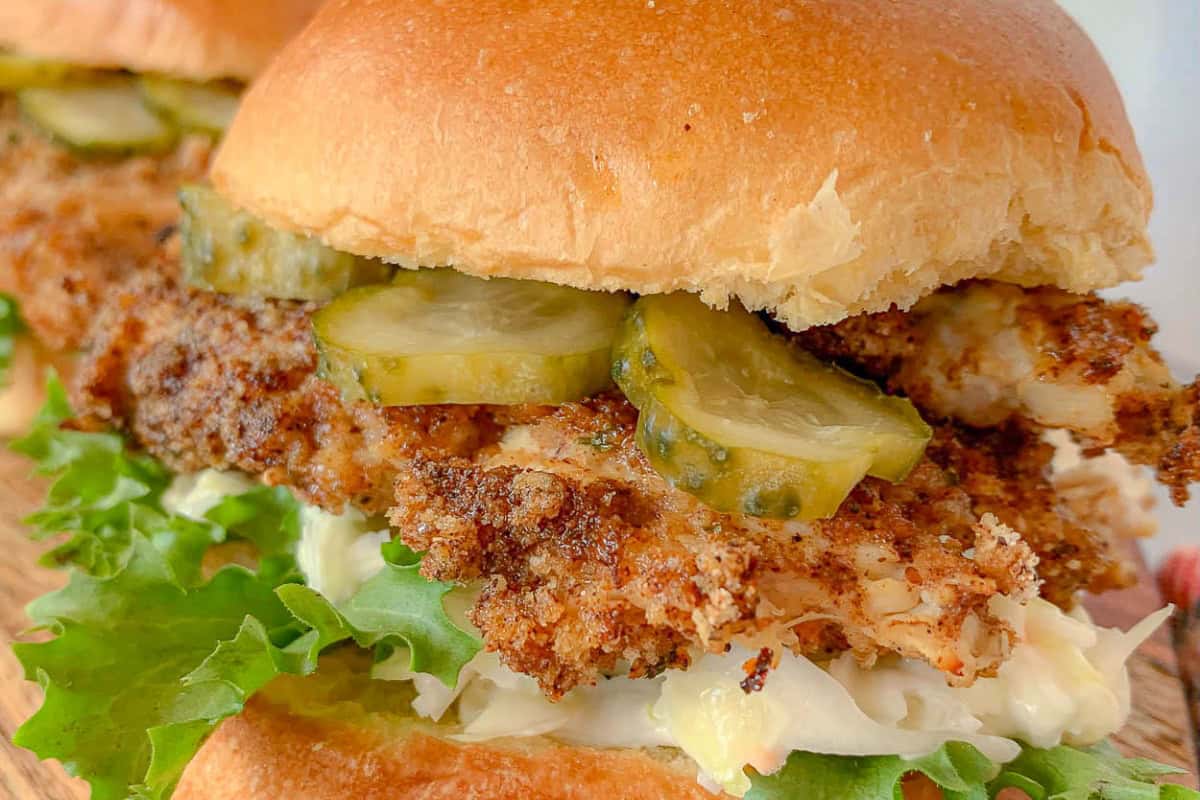 Close-up of a crispy fried chicken sandwich with pickles, lettuce, and creamy coleslaw inside a soft, lightly toasted bun. The sandwich sits on a wooden surface, highlighting its rich textures and mouth-watering appeal.