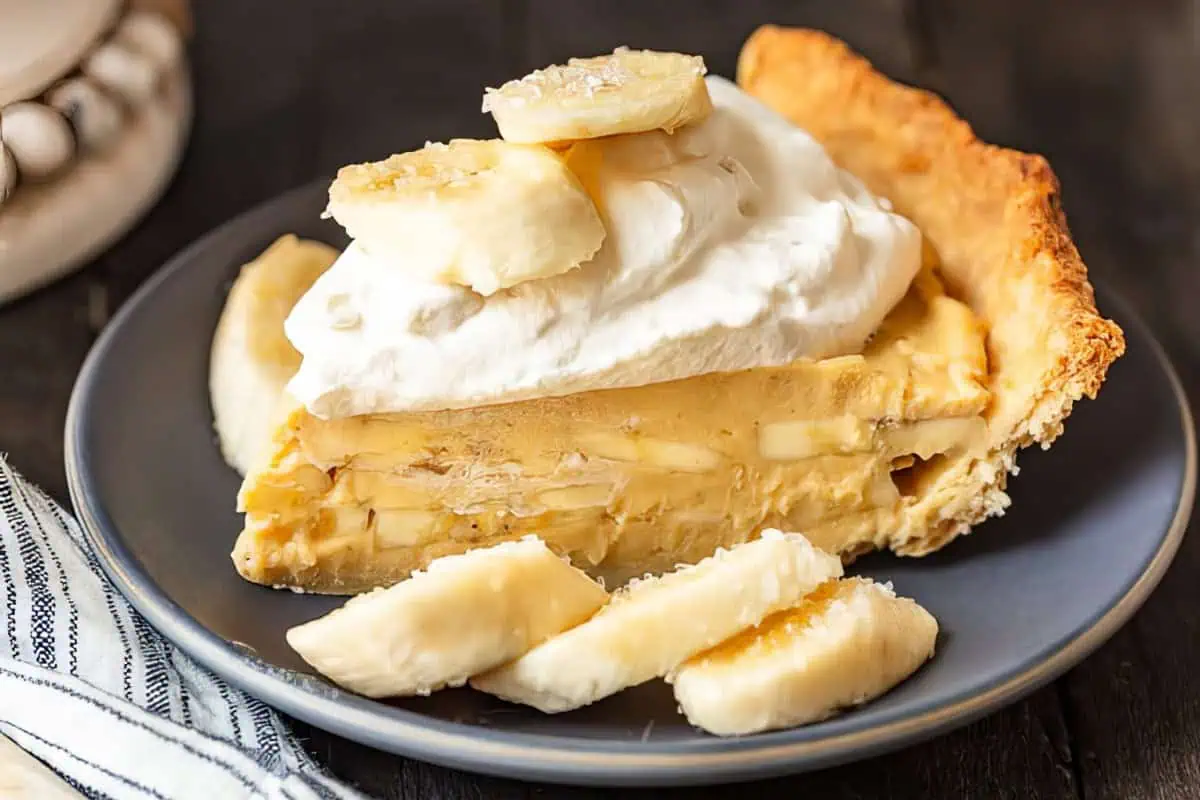 A slice of creamy banana pie topped with a dollop of whipped cream and banana slices is served on a dark plate. More banana slices are placed around the pie slice. The pie has a golden-brown crust, and a striped cloth napkin is visible beside the plate.