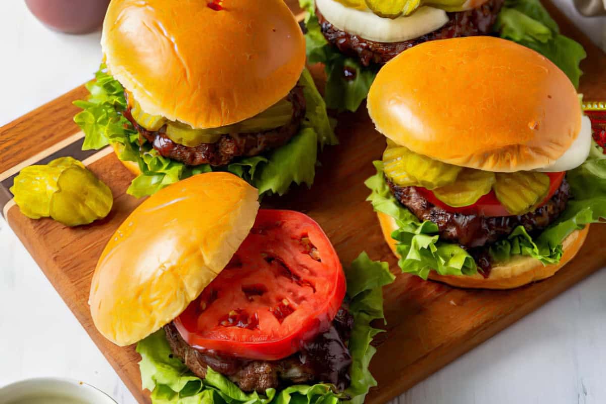 Four juicy burgers with lettuce, tomatoes, pickles, and onions on a wooden board. Toasted buns top the burgers, while one has the bun moved to show the toppings. A few pickle slices are scattered around.