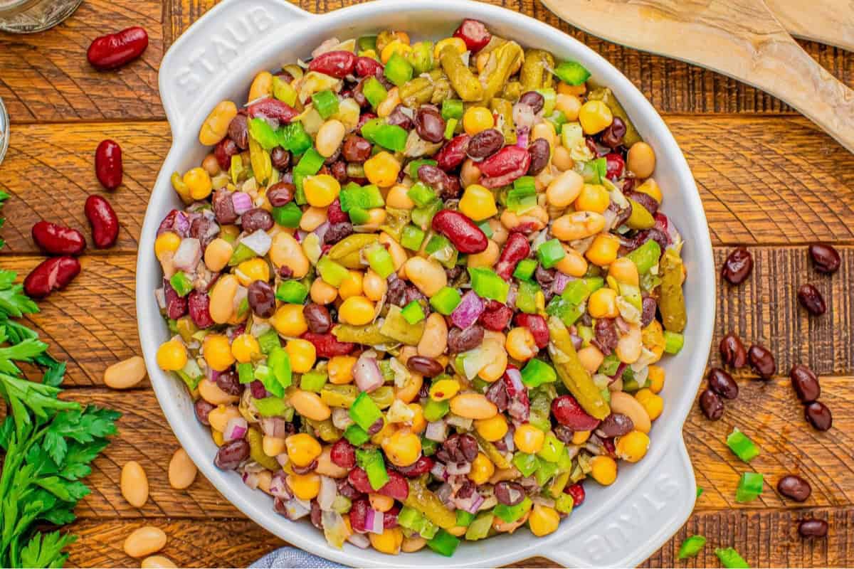 A bowl filled with a colorful bean salad placed on a wooden surface. The salad includes kidney beans, white beans, green bell peppers, corn, red onions, and diced green beans. Parsley sprigs and loose beans are scattered around the bowl.