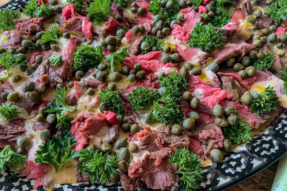 A platter of sliced rare beef topped with fresh green parsley, capers, and a light drizzle of creamy sauce.