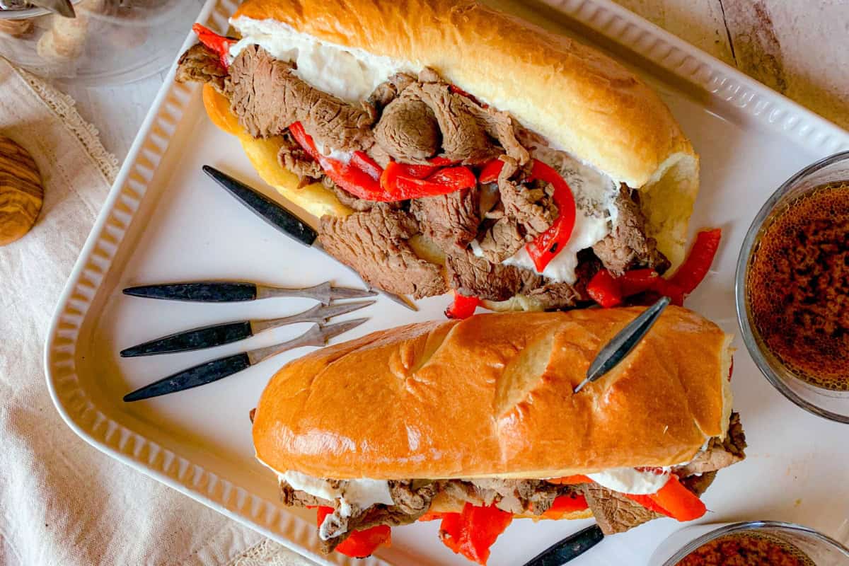 Two Italian beef sandwiches topped with sliced roast beef, red bell peppers, and a white creamy sauce are displayed on a rectangular white plate. Surrounding the sandwiches are small black handled forks and a bowl filled with spices.