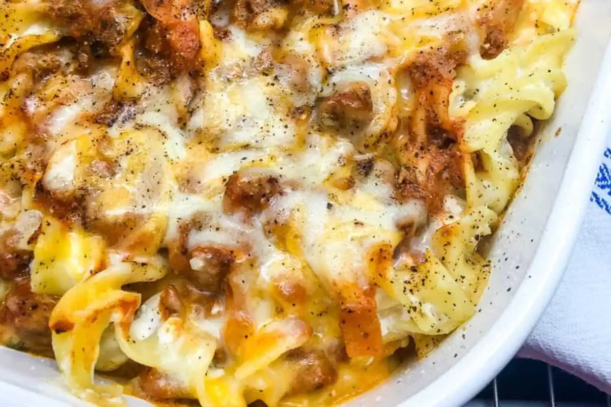 A close-up of a cheesy casserole dish filled with cooked egg noodles, ground meat, and melted cheese. The top is sprinkled with black pepper, and the edges of the dish are slightly browned from baking. The casserole is presented in a white baking dish.