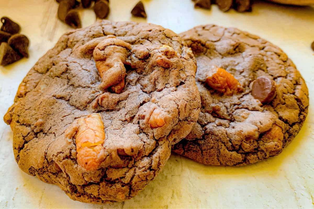 Two thick, chewy double chocolate chip cookies with chunks of chocolate and nuts sit on a light-colored surface. Scattered chocolate chips are visible in the background.
