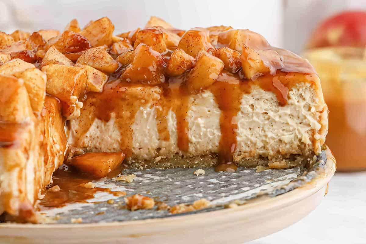 Close-up of a partially sliced caramel apple cheesecake on a plate. The cheesecake has a crumbly crust, a creamy filling, and is topped with caramel sauce and chunks of spiced apples. The background is slightly blurred with hints of more apples and sauce.