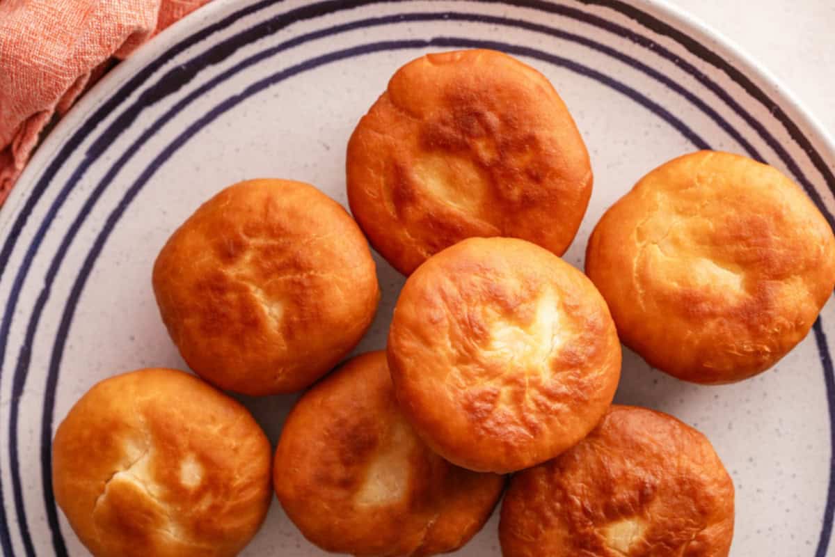 Seven golden-brown, fried dough balls are arranged on a white plate with blue concentric circles. A coral-colored cloth is visible in the corner. The dough balls have a slightly crisp exterior, indicating they were freshly fried.