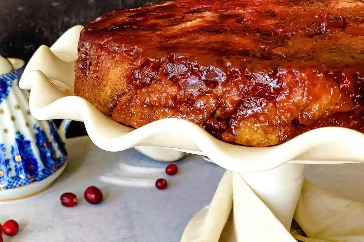 Close-up of an upside-down cranberry cake on a scalloped-edge white cake stand. The cake has a caramelized top with visible cranberries. Nearby are a few scattered cranberries and a blue-and-white teapot in the background.