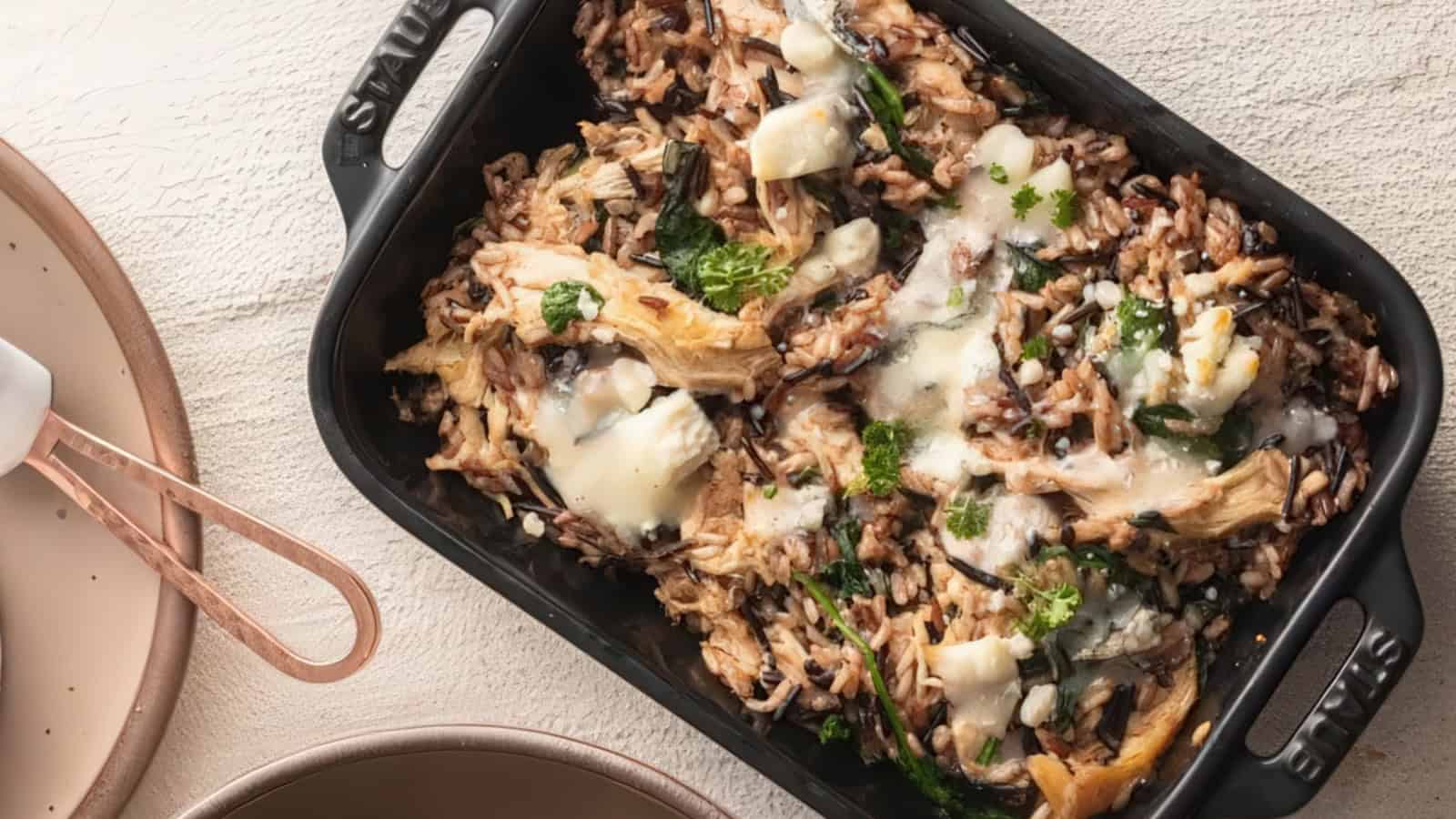 A black baking dish filled with a creamy mushroom and rice casserole topped with herbs and melted cheese. The dish is set on a textured white surface, with two empty beige plates and a copper-handled serving spatula nearby.