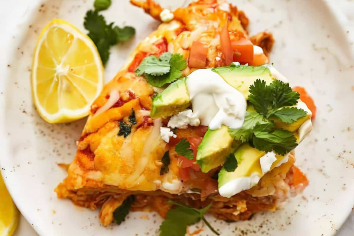 A close-up of a plate with a serving of enchilada dish, topped with melted cheese, diced tomatoes, avocado slices, sour cream, and cilantro. A lemon wedge is on the side, with garnish scattered around the plate.