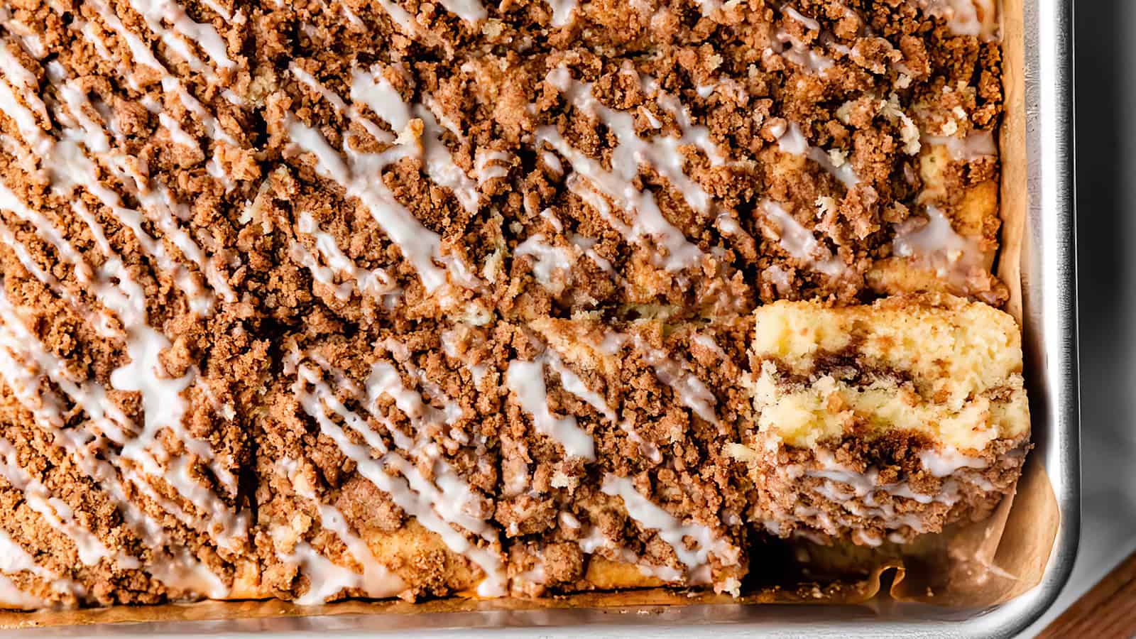 A close-up view of a crumb coffee cake in a rectangular baking dish, topped with a generous layer of crumbled streusel and a drizzle of white icing. One piece is cut out from a corner, revealing the cake's moist texture and layers.