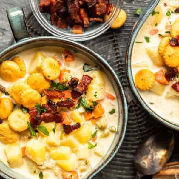 Two bowls of creamy soup topped with crispy bacon bits, oyster crackers, and chopped herbs. The soup contains chunks of potato and diced vegetables. A small bowl filled with extra crispy bacon bits and a green napkin are visible alongside the bowls. Two spoons rest nearby.