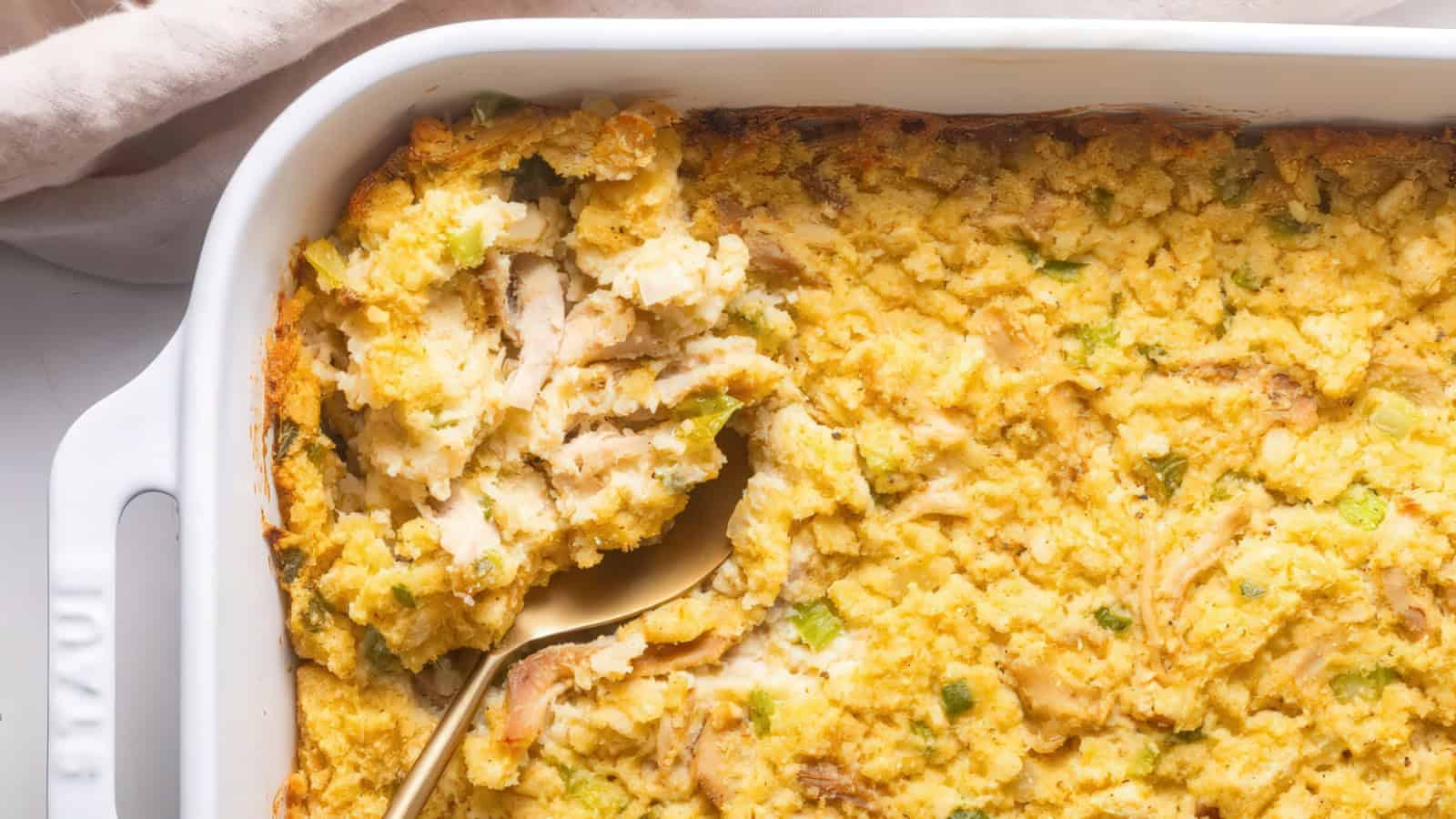 A close-up image of a golden-brown casserole in a white baking dish. The dish appears to contain a bread-based stuffing mixed with visible pieces of celery and herbs. A serving spoon is partially scooping out a portion.