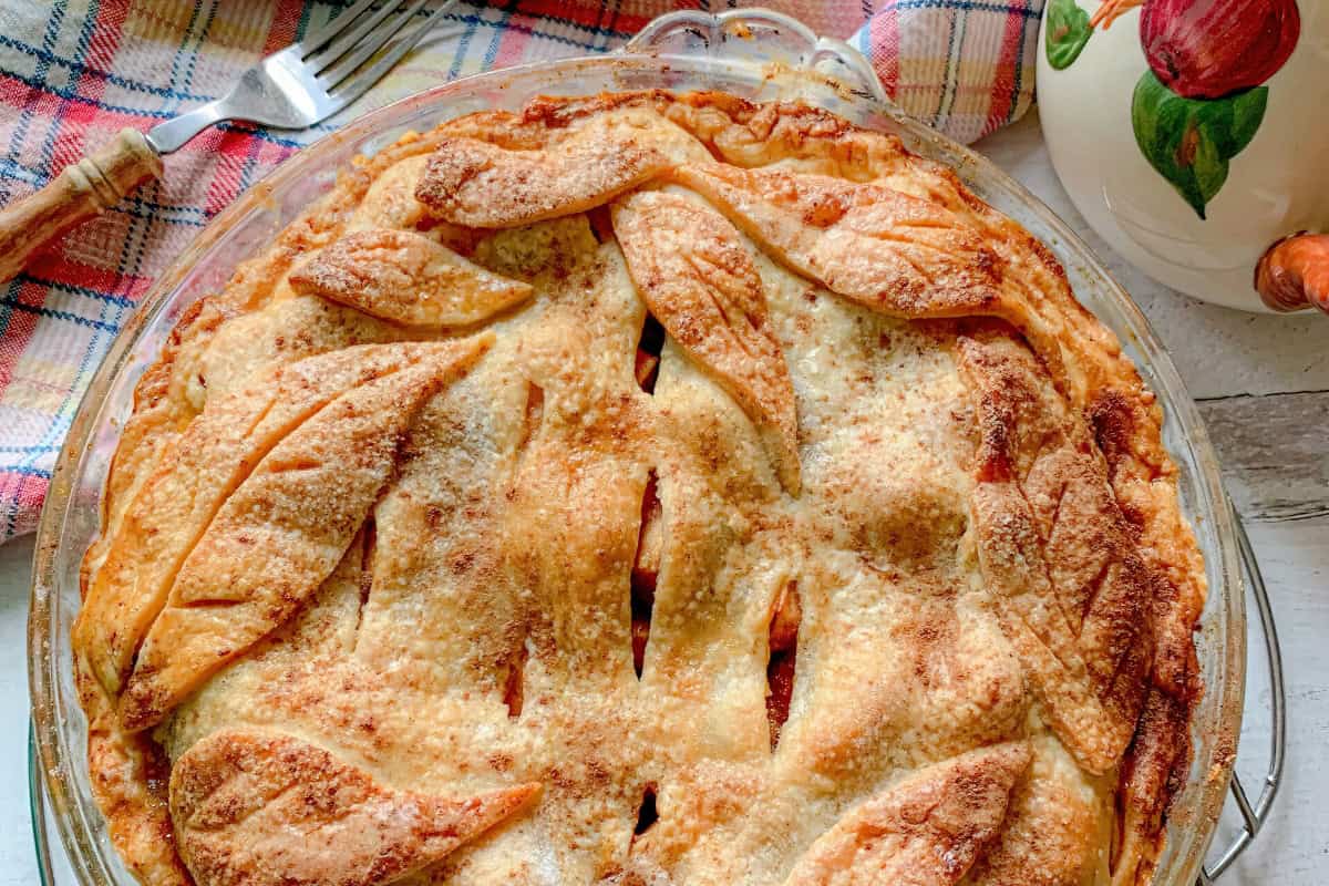 A freshly baked pie with a golden, flaky crust decorated with leaf-shaped patterns sits on a table. A fork rests on a colorful plaid napkin beside the pie, and a ceramic pitcher with a fruit design is partially visible.