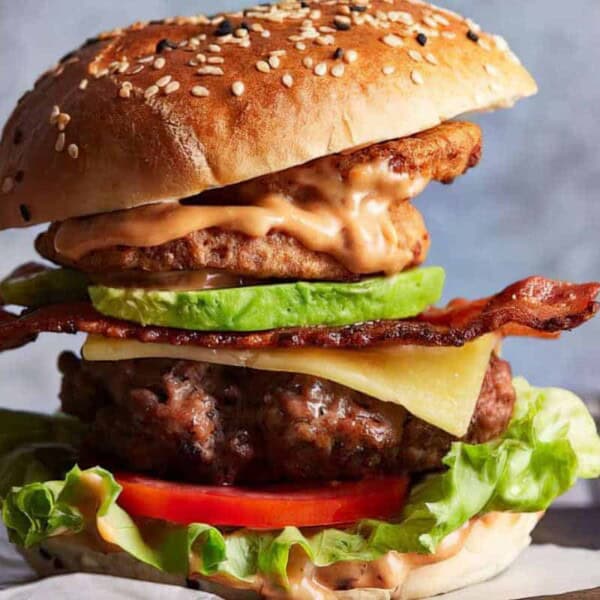A towering hamburger with a sesame seed bun, featuring layers of lettuce, tomato, a thick beef patty, melted cheese, crispy bacon strips, avocado slices, a fried onion ring, and drizzled with sauce. The background is softly blurred, focusing on the burger.