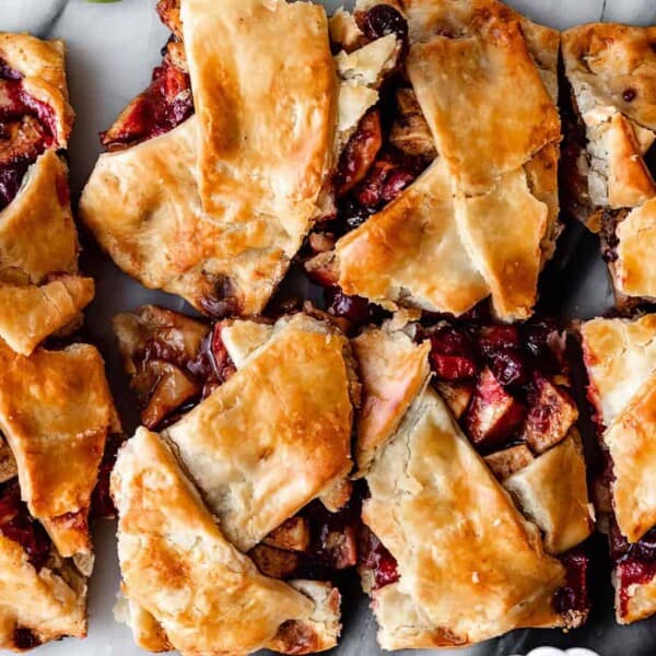 A flaky, golden-brown pastry is divided into nine irregularly cut squares. It is filled with a mixture of baked apples and cranberries, with some fruit juices visible, creating a rustic, homemade appearance. The pastry is displayed on a white marble surface.