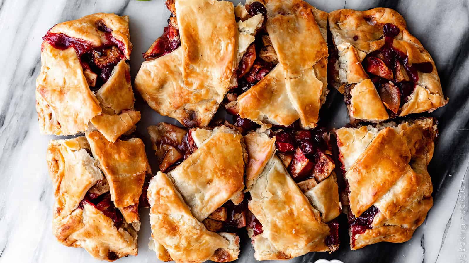 A flaky, golden-brown pastry is divided into nine irregularly cut squares. It is filled with a mixture of baked apples and cranberries, with some fruit juices visible, creating a rustic, homemade appearance. The pastry is displayed on a white marble surface.
