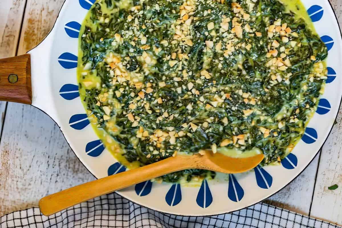 A colorful skillet filled with creamy spinach topped with chopped nuts. The skillet has a wooden handle and a blue pattern around the edge. A wooden spoon rests inside the dish. The skillet is placed on a rustic wooden surface with a checkered cloth nearby.
