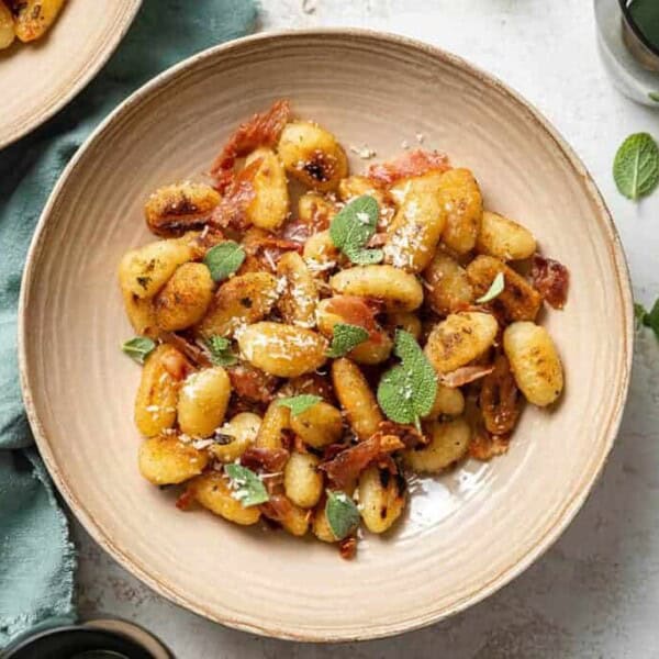 A beige bowl of cooked gnocchi topped with crispy prosciutto, grated cheese, and sage on a light surface with a green napkin.