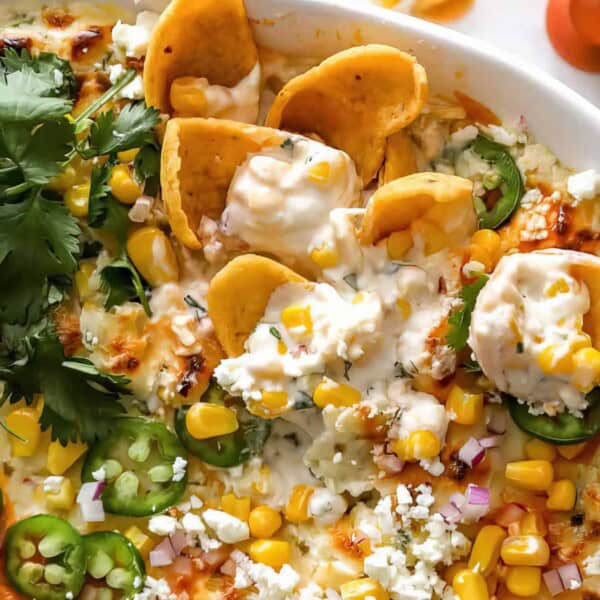 A close-up of a dish featuring cheesy nachos topped with corn, jalapeño slices, crumbled cheese, fresh cilantro, and dollops of creamy sauce. The colorful toppings are spread out on a crispy layer of baked cheese, creating a vibrant and appetizing presentation.