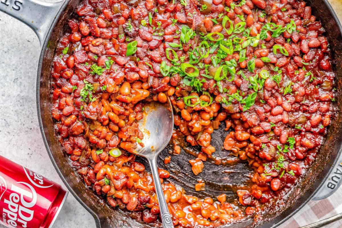 A cast iron skillet filled with baked beans, garnished with chopped green onions and herbs. A spoon rests in the skillet, and a portion of beans has been served. A can of Dr Pepper soda lies nearby.
