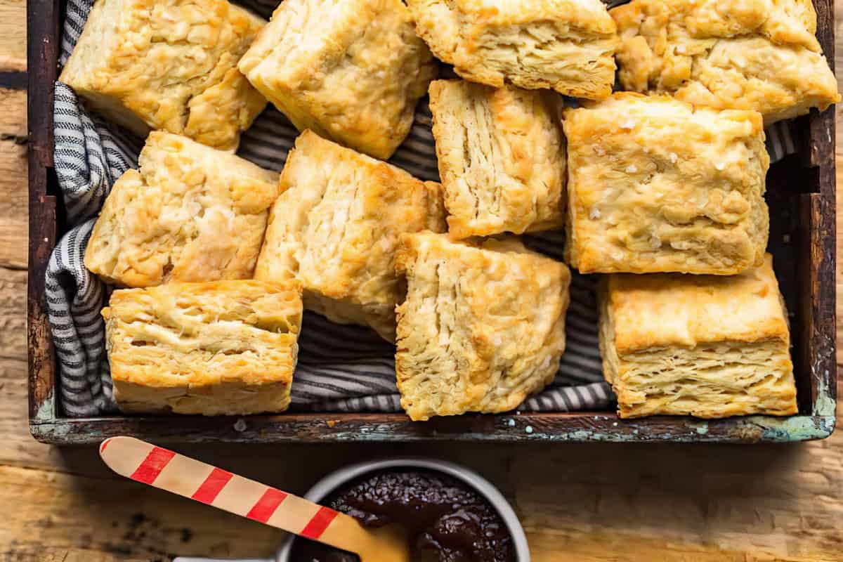 A wooden tray filled with several golden-brown, flaky biscuits on a striped cloth. In front of the tray is a small bowl containing a dark-colored spread and a red-striped wooden spreader resting on its edge. The scene is set on a rustic wooden surface.