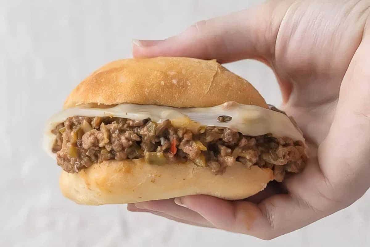 A hand holding a sandwich filled with ground beef mixed with vegetables, topped with a slice of melted cheese, and encased in a soft bun. The background is out of focus, highlighting the sandwich.