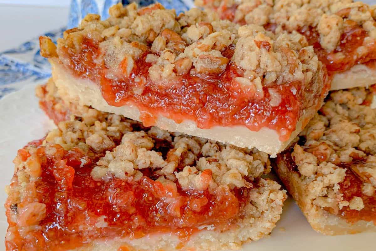 Close-up of a stack of strawberry crumble bars resting on a white surface. The bars have a crumbly oat topping and a thick layer of vibrant red strawberry filling over a buttery base. The texture appears crisp and delicious. Blue and white patterned fabric is partially visible in the background.