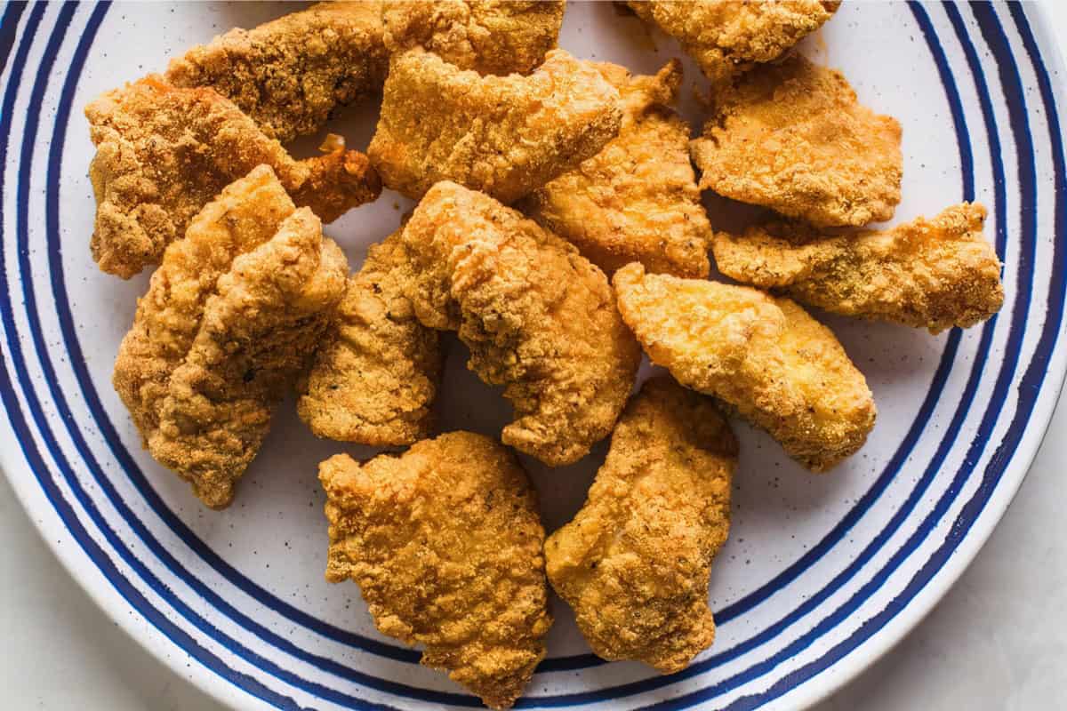 A white plate with blue stripes holds a serving of golden-brown fried catfish nuggets, arranged in a circular pattern. The nuggets appear crispy and well-seasoned. The background is a clean white surface.