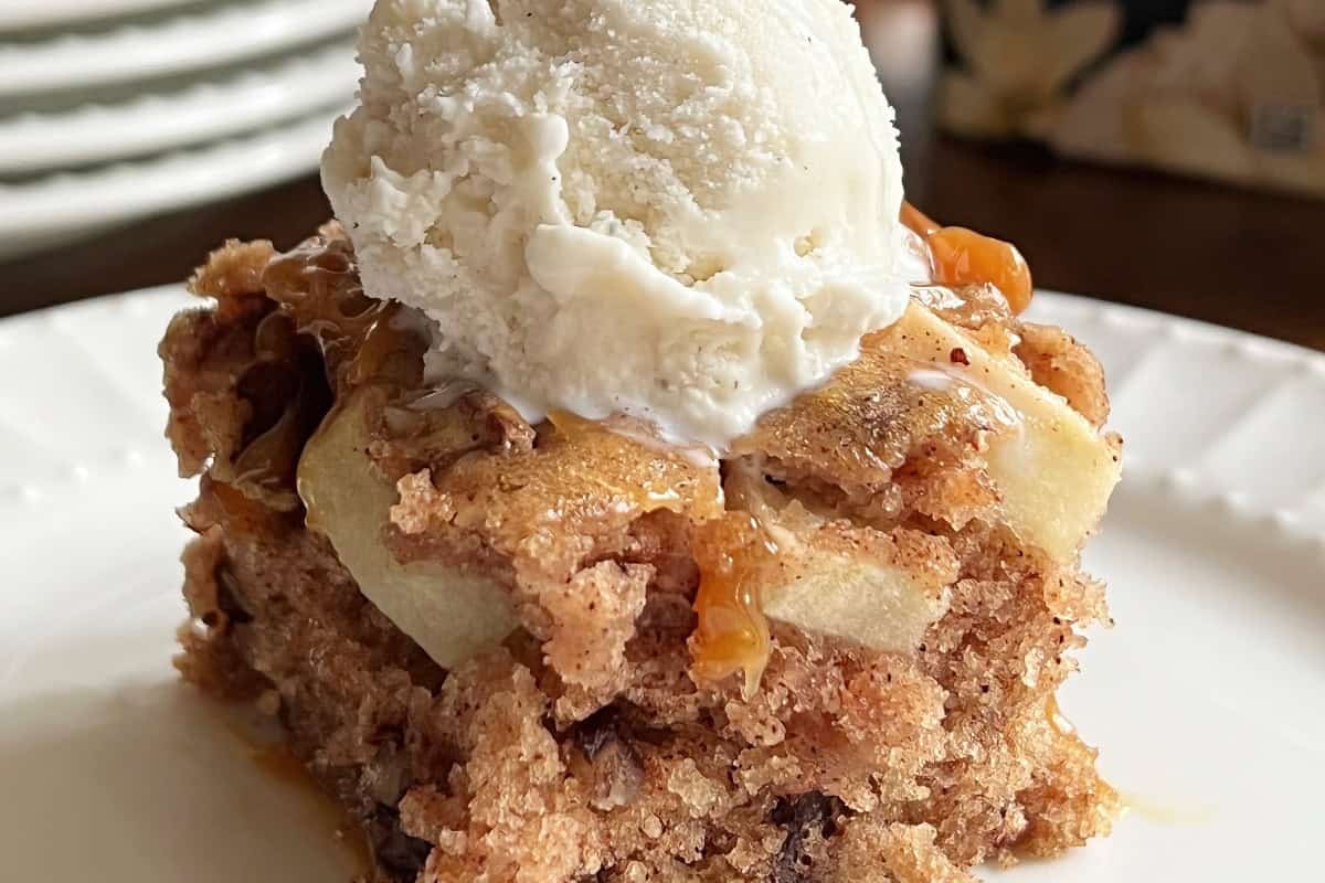 A close-up of a dessert square on a white plate, featuring a moist, spiced apple cake topped with a scoop of vanilla ice cream and drizzled with caramel sauce. The cake has visible apple chunks and a soft, crumbly texture.