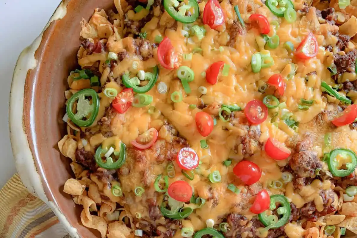 A close-up of a dish featuring a layer of melted cheese, ground meat, sliced jalapeños, halved cherry tomatoes, and chopped green onions on top of corn chips. The dish appears to be a flavorful and colorful casserole or nacho platter.