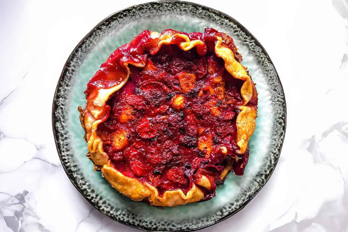 A rustic red berry crostata with a crispy, golden-brown edge is placed on a green ceramic plate. The crostata has a flaky crust with a filling of mixed berries, visibly baked to a juicy, slightly caramelized finish. The background is a white marble surface.