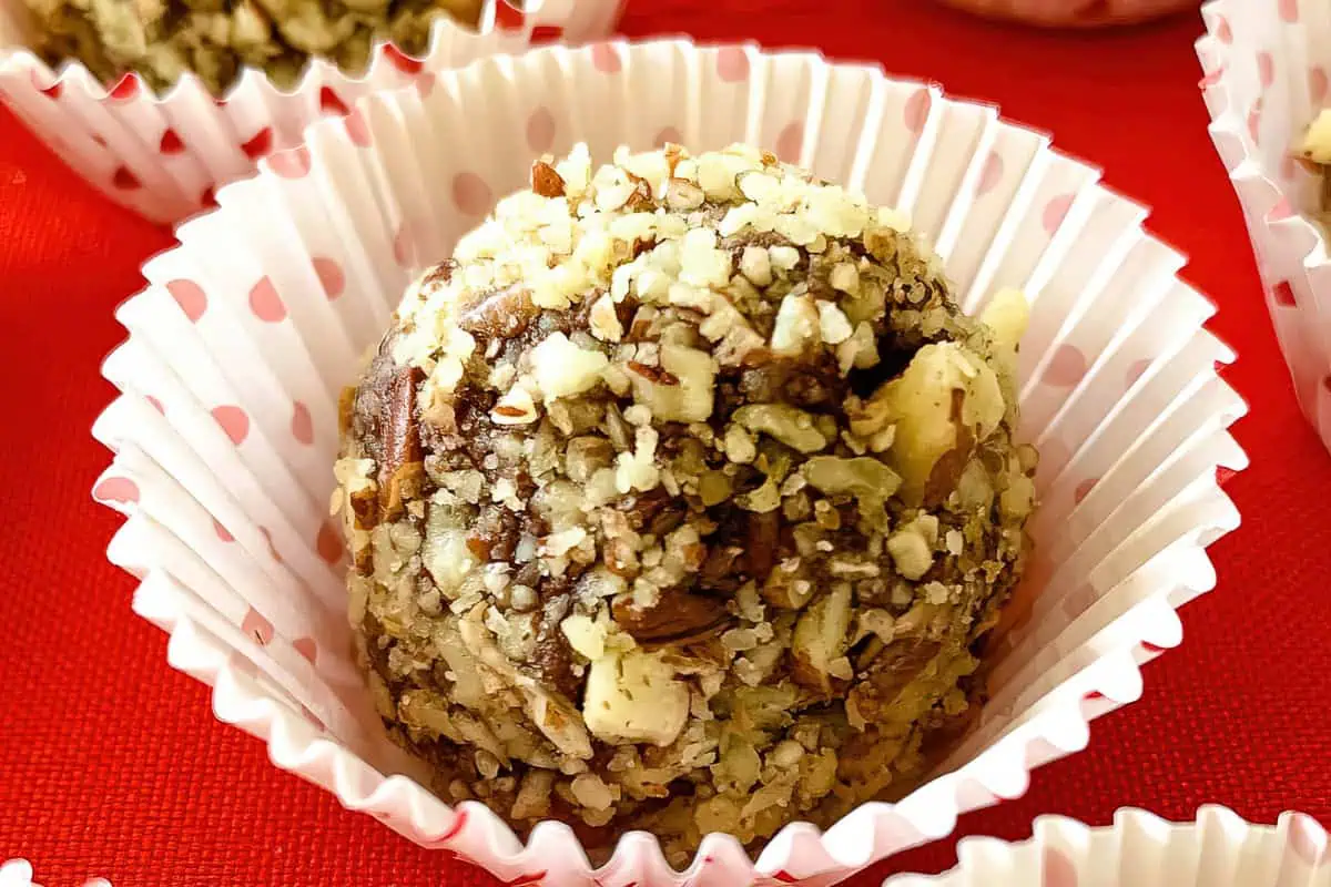 Close-up of a dessert ball coated in chopped nuts and seeds, placed in a white, ruffled paper cup with red polka dots. The background features a red surface and other similar dessert balls in the distance.