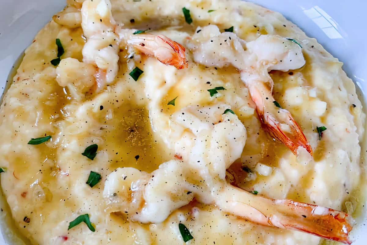 A close-up of a savory dish featuring creamy grits topped with grilled shrimp. The grits are garnished with melted cheese, chopped herbs, and a light drizzle of sauce. The dish appears rich and appetizing, served on a white plate.
