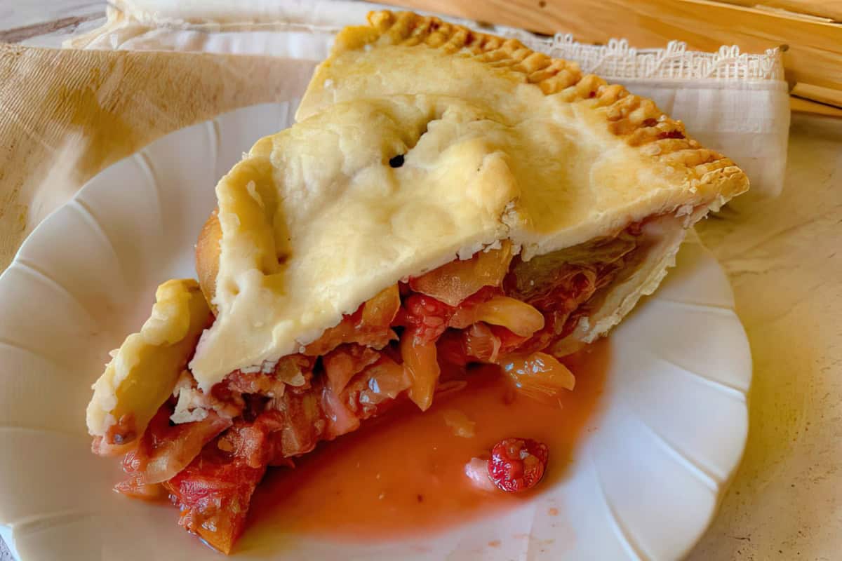 A slice of homemade apple pie on a white plate. The pie has a golden, flaky crust, with pieces of apple and a rich, slightly gooey filling visible. There is a small amount of sauce pooling on the plate. The background includes a light cloth and a wooden surface.