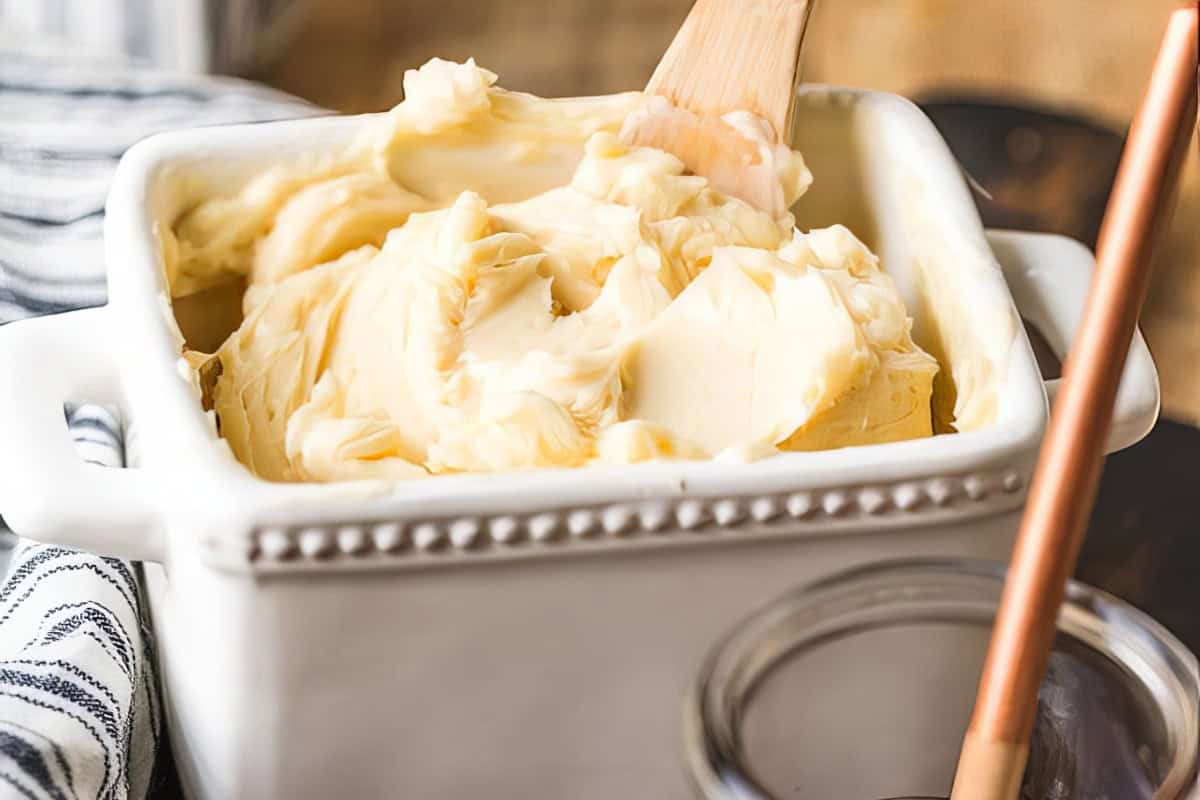 A white ceramic dish is filled with creamy butter, which is being smoothed by a wooden spatula. A striped cloth is visible to the left, and a blurred background suggests a kitchen setting. The edges of a mason jar and a spoon handle are partially visible on the right.