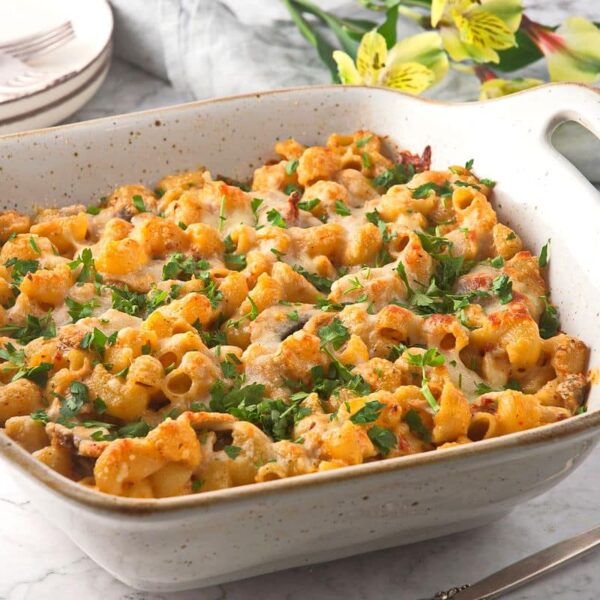 A white ceramic baking dish filled with baked pasta, topped with melted cheese and garnished with chopped parsley, sits on a marble countertop.