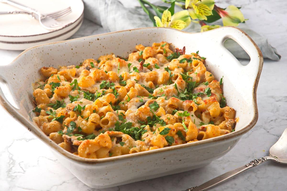 A white ceramic baking dish filled with baked pasta, topped with melted cheese and garnished with chopped parsley, sits on a marble countertop.