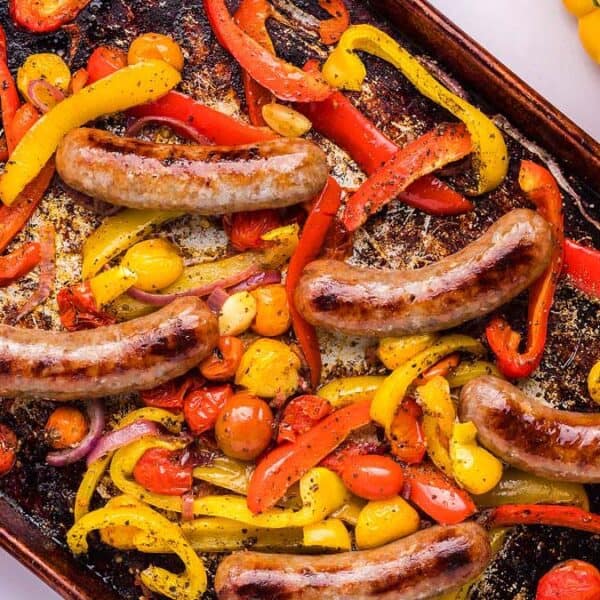 Sausages, sliced red and yellow bell peppers, cherry tomatoes, and garlic cloves arranged on a baking tray.
