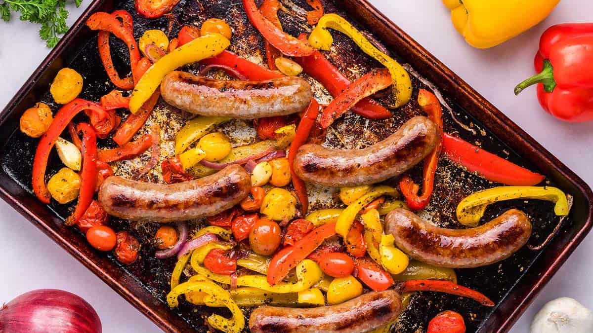 Roasted sausages on a baking tray with sliced red and yellow bell peppers, cherry tomatoes, and garlic cloves.