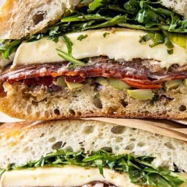 A close-up of two stacked sandwiches with cheese, salami, arugula, tomato, and olives on rustic bread.