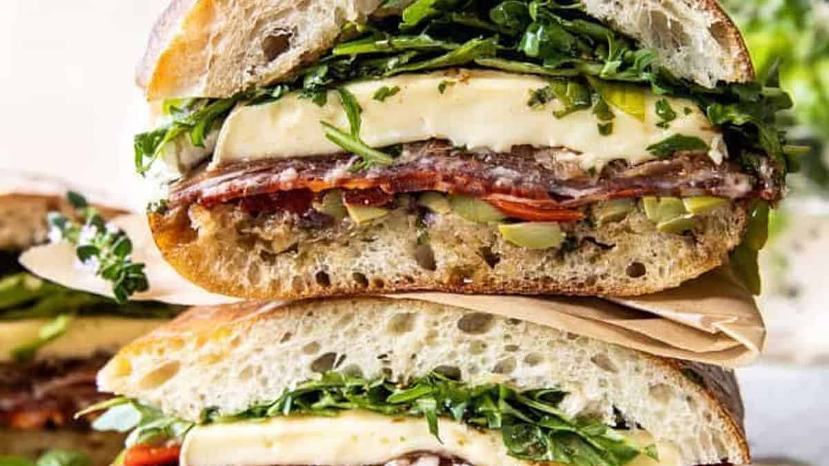 A close-up of two stacked sandwiches with cheese, salami, arugula, tomato, and olives on rustic bread.