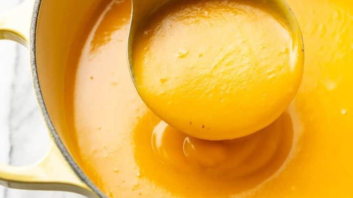 A metal ladle lifts creamy orange soup from a yellow pot.